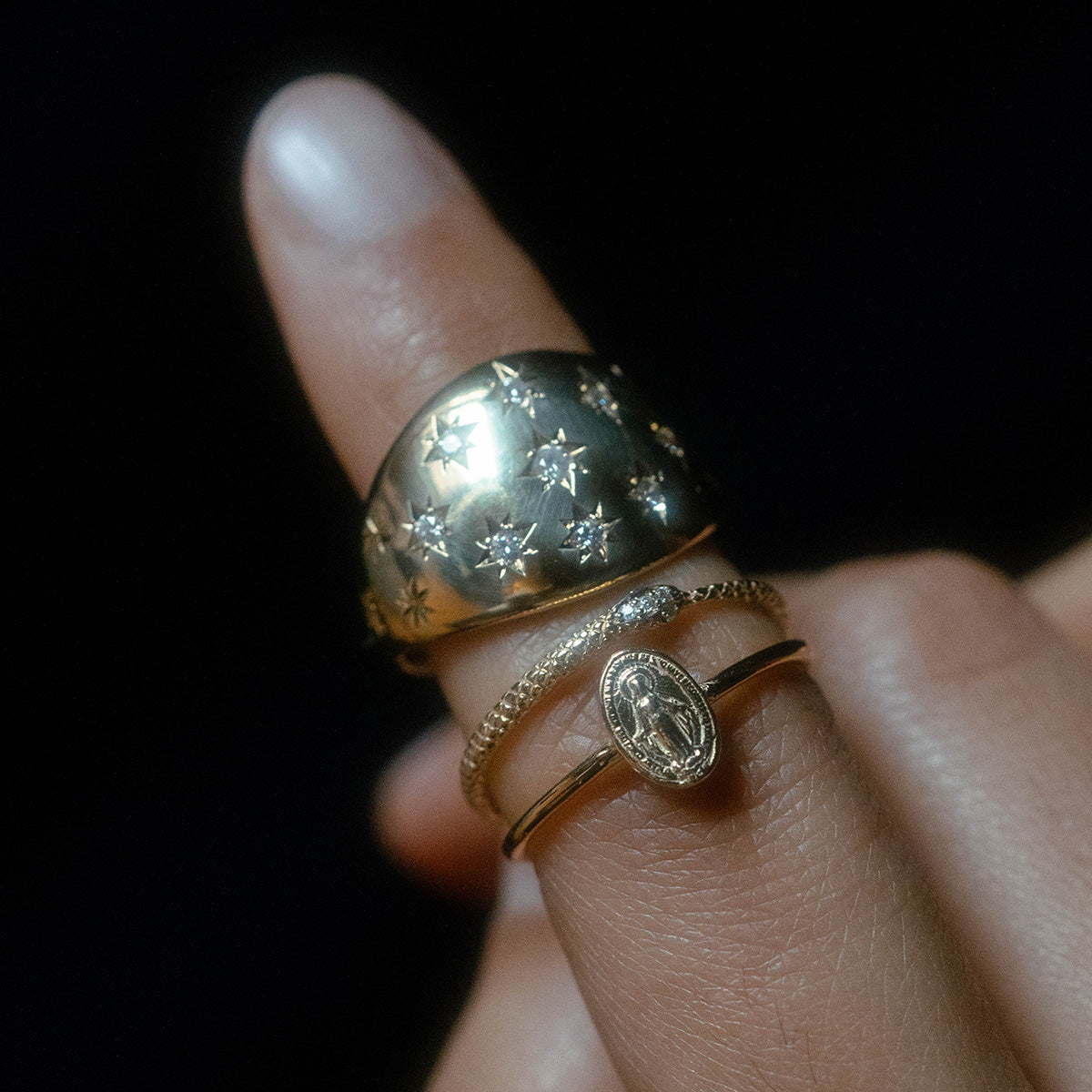 A close-up of a hand wearing three gold rings, including the Bombe Diamond Ring, with a dark background.