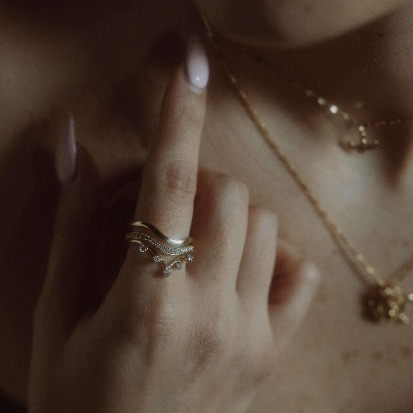 A close-up of a hand with pale pink nails showcases a solid gold ring with small charms and the 14k Low Tide Curved Band, paired with a gold necklace. The softly blurred background adds an elegant touch.