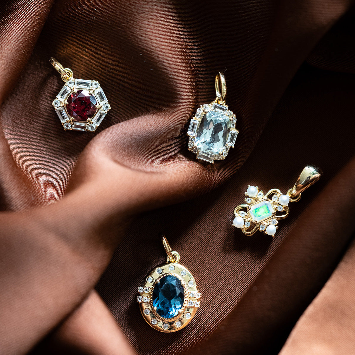 Four Vintage Inspired Pendant Charms with gold vermeil settings rest on brown satin; each features ornate detailing, small clear stones, and vibrant gemstones in red, light blue, dark blue, and green.