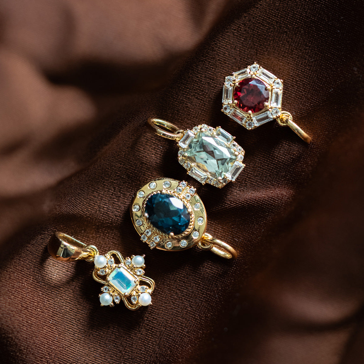 A Vintage Inspired Pendant Charm with ornate gold details, colorful gemstones, and pearl accents rests on rich brown satin, evoking Period Piece jewelry with intricate designs and sparkling blue, green, red, and turquoise stones.