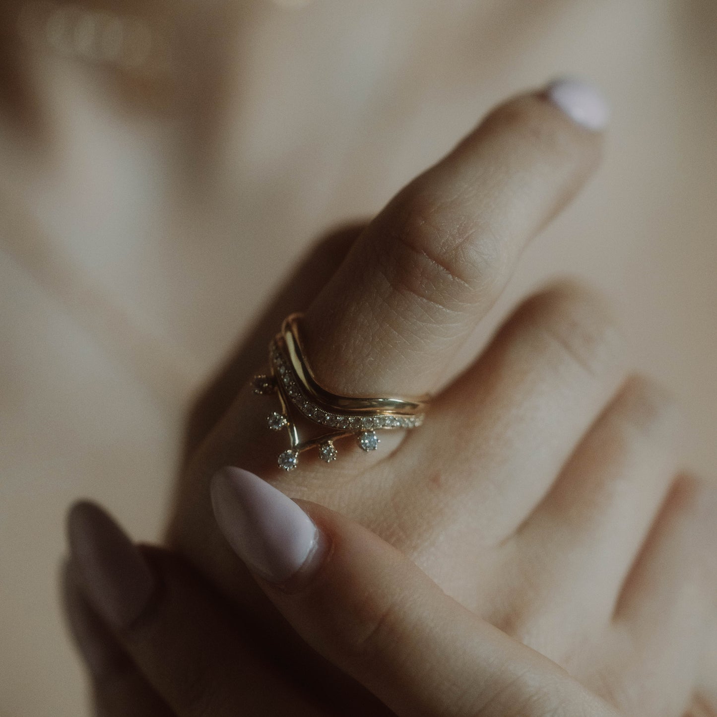 A close-up shows a hand with light pink manicured nails wearing the 14k Low Tide Curved Band stacked with a solid gold ring—one plain, one featuring small dangling gems—on the index finger against a softly blurred background.