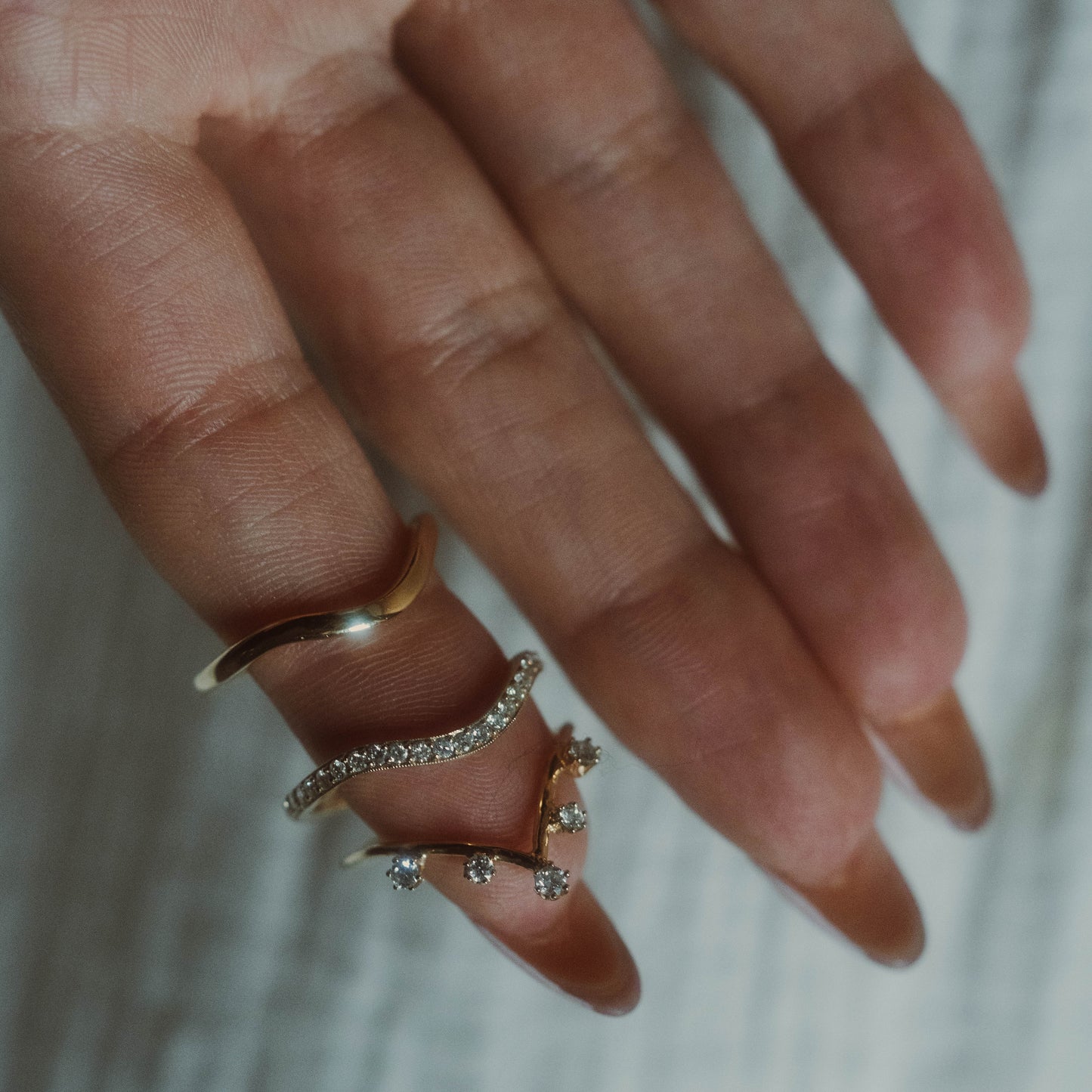 A hand wears three gold rings on the middle finger: a solid band, one with dangling diamond-like stones, and the 14k Low Tide Curved Band. Each stacking ring features a unique wavy design against a softly blurred background.