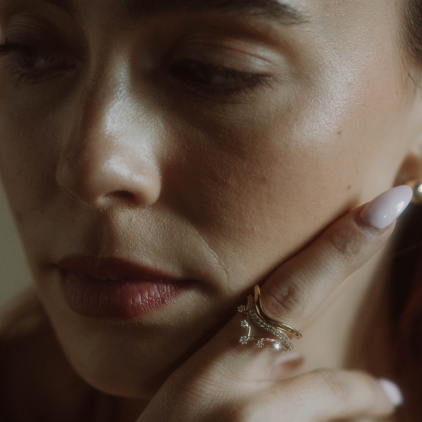 A close-up of a womans face focuses on her lips and cheek as she rests her chin on her hand, showing off the 14k Low Tide Curved Band and pale manicured nails. Soft, natural lighting enhances the texture of her skin.