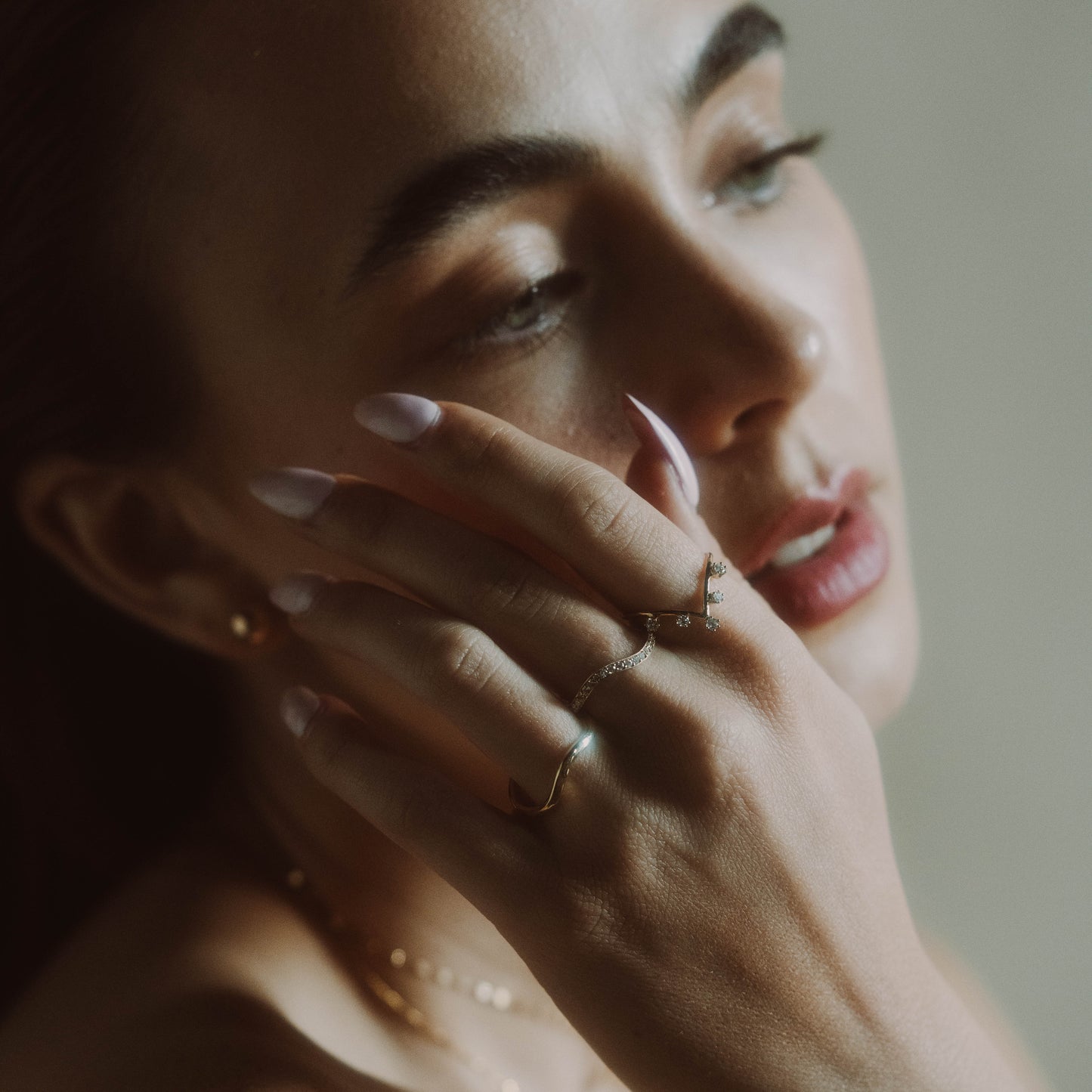 A woman with manicured nails touches her face, highlighting the 14k Low Tide Curved Band and delicate rings. She wears soft makeup, while the image features a blurred effect and neutral tones.