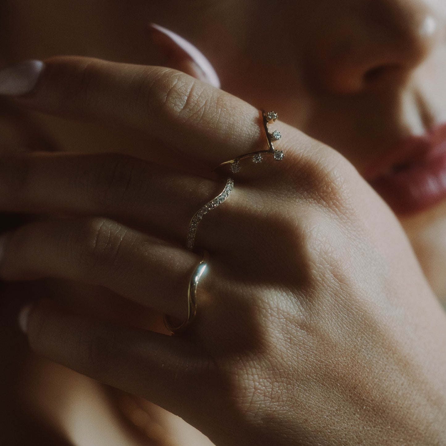 A close-up of a manicured hand wearing the 14k Low Tide Curved Band and two gold rings with delicate designs and small stones, held near the lips against a softly blurred background.