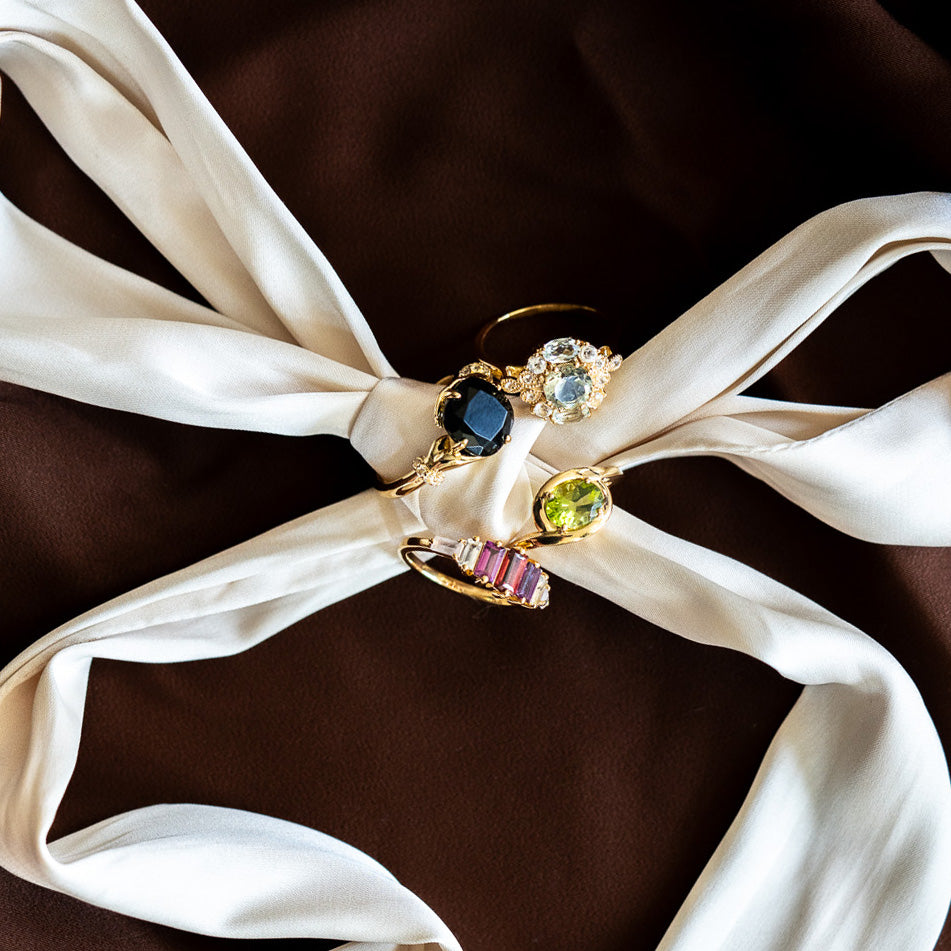 The Peridot Poetry Statement Ring and three other 14k gold vermeil gemstone rings are displayed on a twisted white silk ribbon against a brown fabric background.