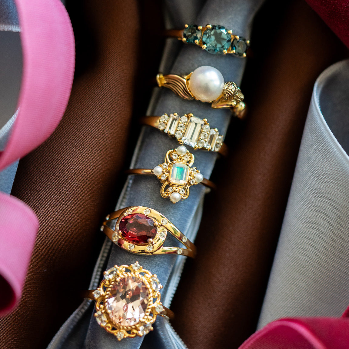 Five ornate rings, including the Royal Blush Cocktail Ring, showcase gemstones in green, pearl, clear, red, and pale pink on a gray holder with pink and blue satin ribbons set against brown fabric.