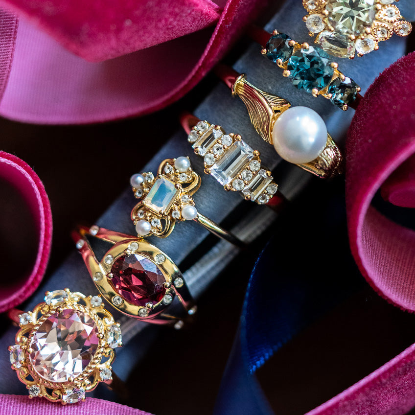 A close-up of five ornate rings, including the Royal Blush Cocktail Ring in 14k gold vermeil with colorful gemstones and diamonds, displayed on a navy blue velvet holder surrounded by pink velvet ribbons.