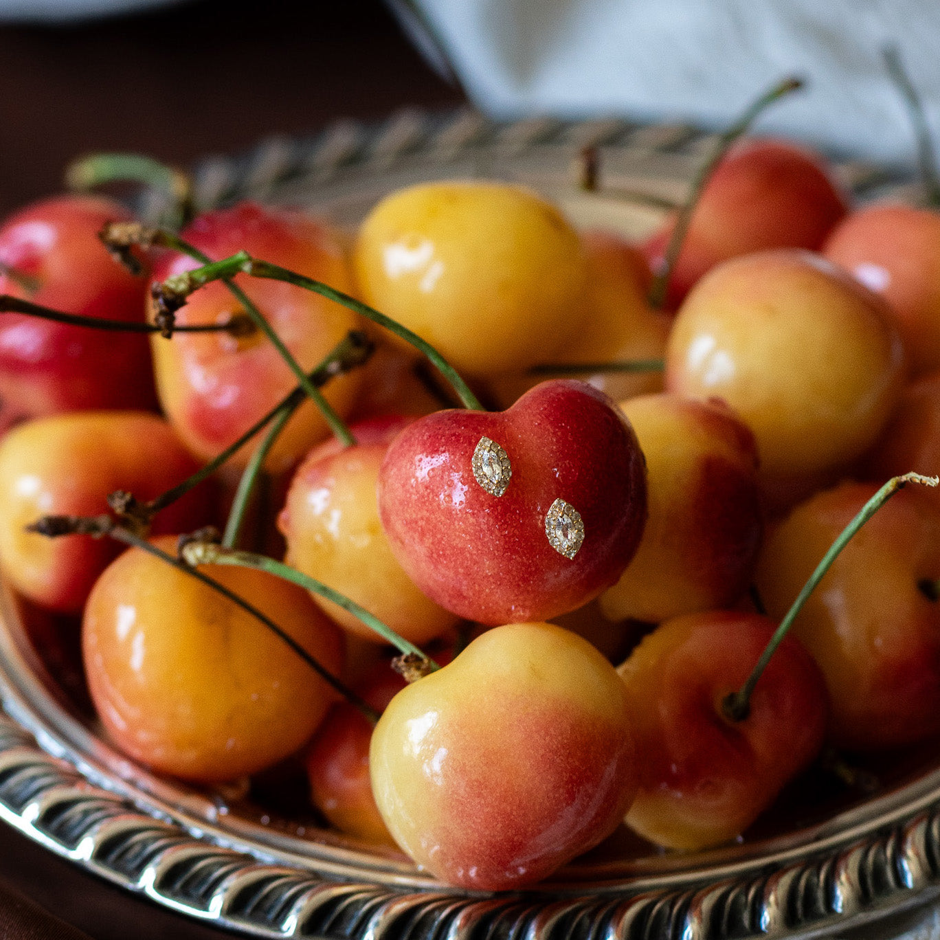 A close-up of a silver tray filled with vibrant Rainier cherries, their yellow and red hues with green stems shining. In the foreground, a cherry is adorned with two Green Amethyst Marquise Muse Studs, reminiscent of vintage earrings.