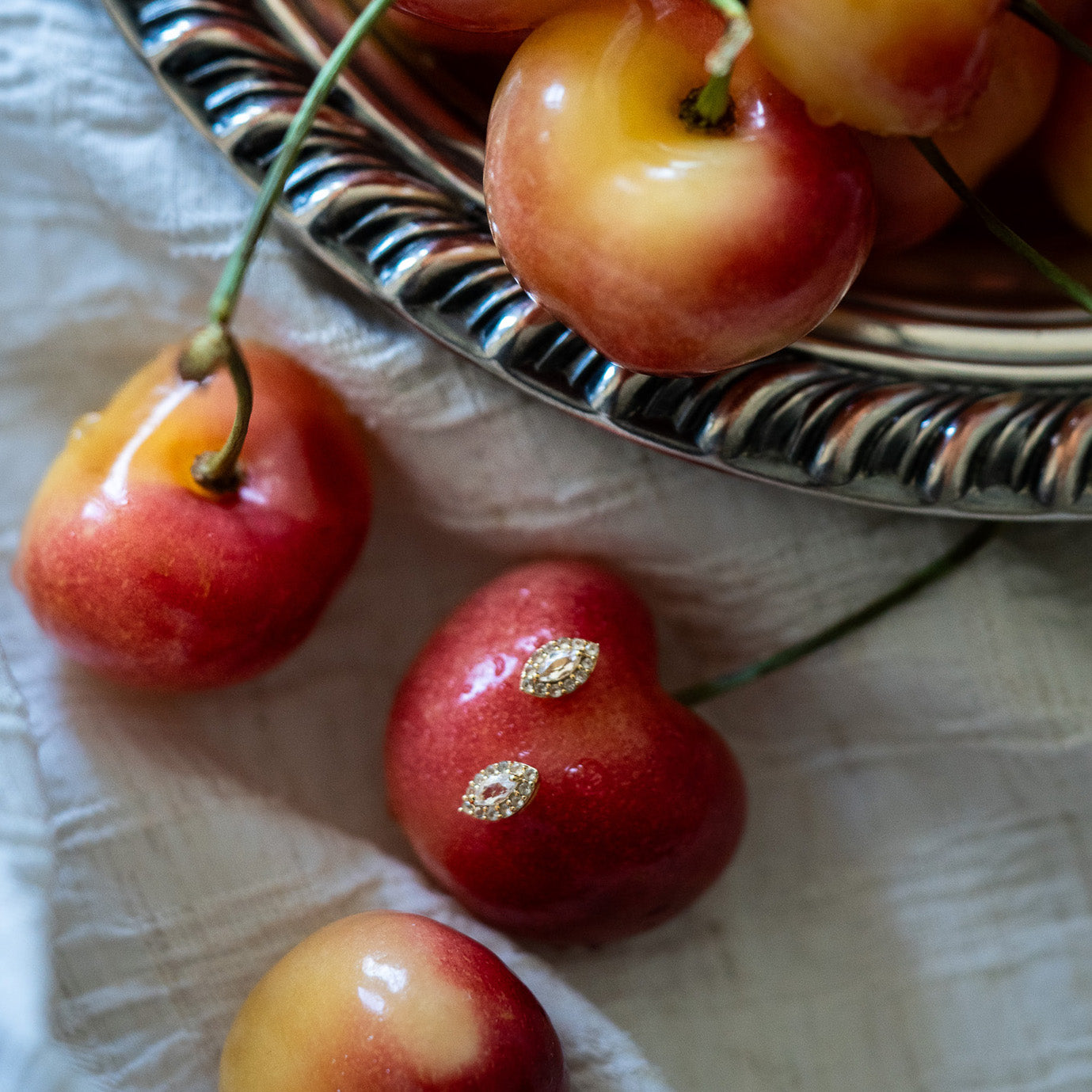 A close-up of Rainier cherries on a white cloth, with the Green Amethyst Marquise Muse Studs—leaf-shaped gold earrings—resting on one of the cherries near a silver dish.