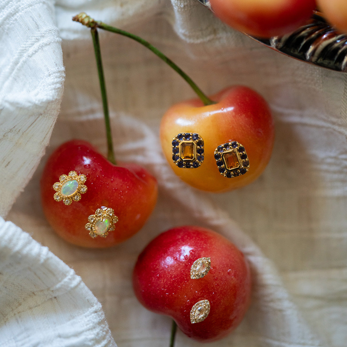 Three cherries rest on a white cloth, each adorned with Opal Majesty Studs, featuring Ethiopian opals and intricate designs. A metal tray filled with more cherries is visible in the top right corner.