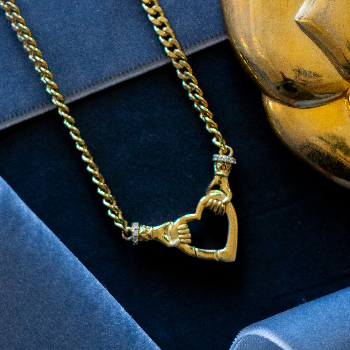 The Hand Over Heart Charm Holder Necklace, crafted in 14k gold vermeil and featuring a pendant of two hands forming a heart, is displayed on dark blue velvet beside a gold decorative piece.