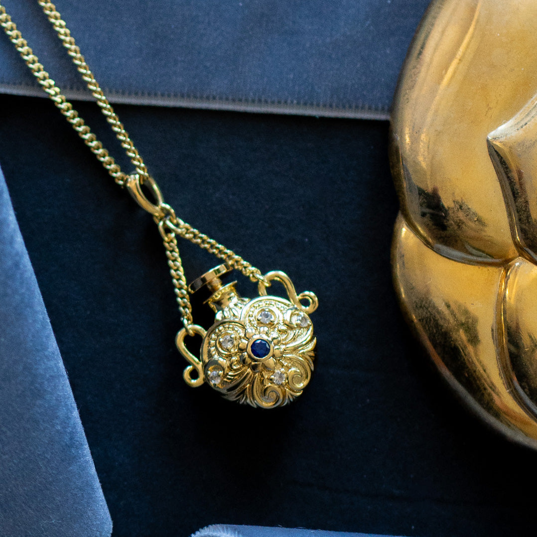 The Juliet Vial Pendant Necklace is a vintage-inspired gold piece with ornate floral designs and a small blue gemstone at its center, displayed next to a shiny gold decorative object on a dark surface.