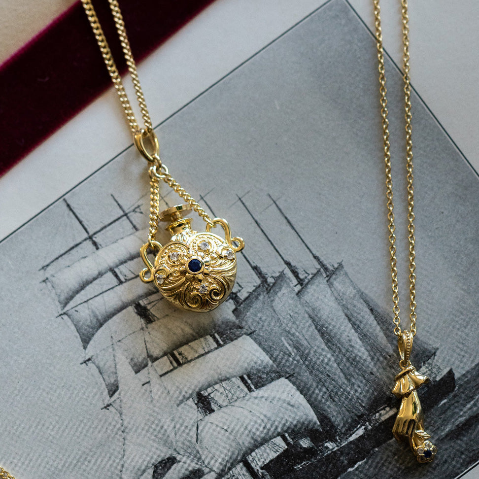 The Juliet Vial Pendant Necklace, shaped like a hand holding an object, is paired with a vintage-inspired gold necklace featuring filigree and a blue stone, both resting on a sepia-toned photo of a sailing ship.