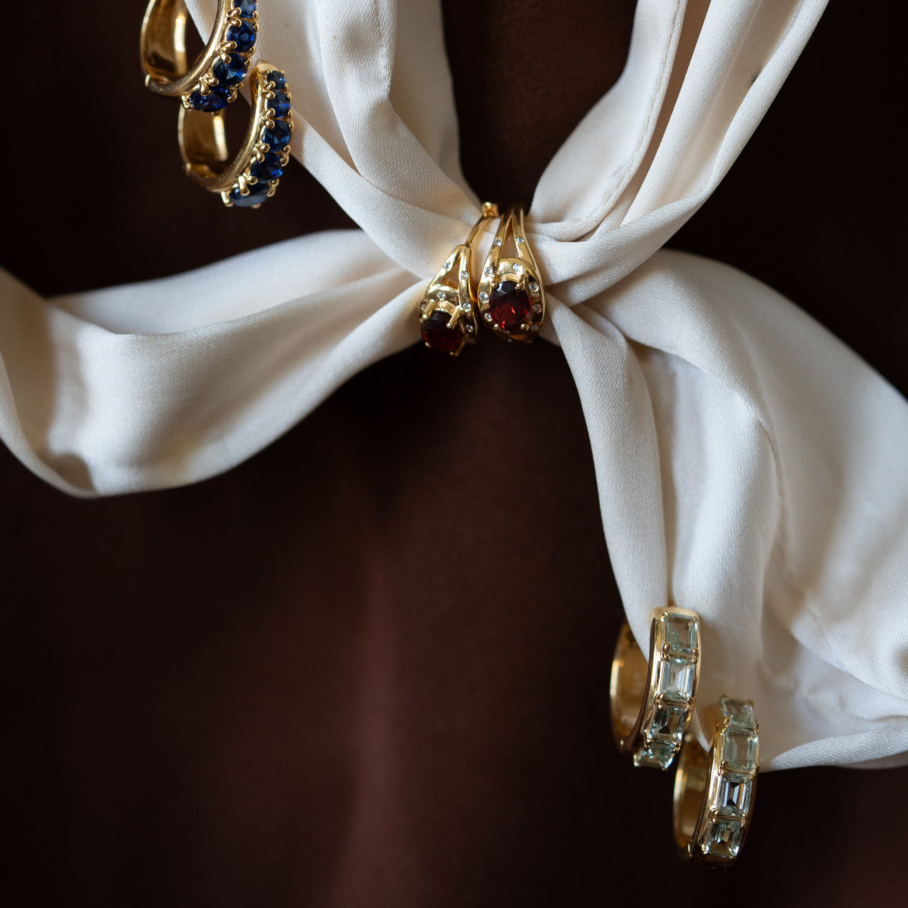 Three pairs of gold earrings, including elegant Green Amethyst Era Huggie Hoops, are arranged on a tied white cloth against a dark brown background—a scene with vintage charm.
