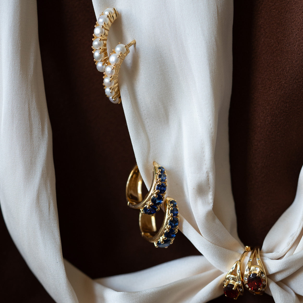 Nonnas Pearl Hoop Earrings—a set of three gold vermeil hoops, one with freshwater pearls, one with blue stones, and one with red stones—are arranged on white silk fabric against a brown background.