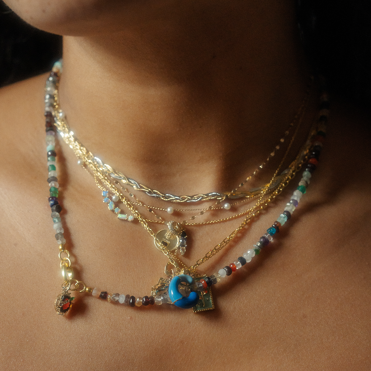 A close-up of a neck adorned with layered necklaces, featuring the Shades of You Ombre Birthstone Necklace with multicolored beads and gold chains. Pendants include a blue charm and gold accents against a softly blurred background.