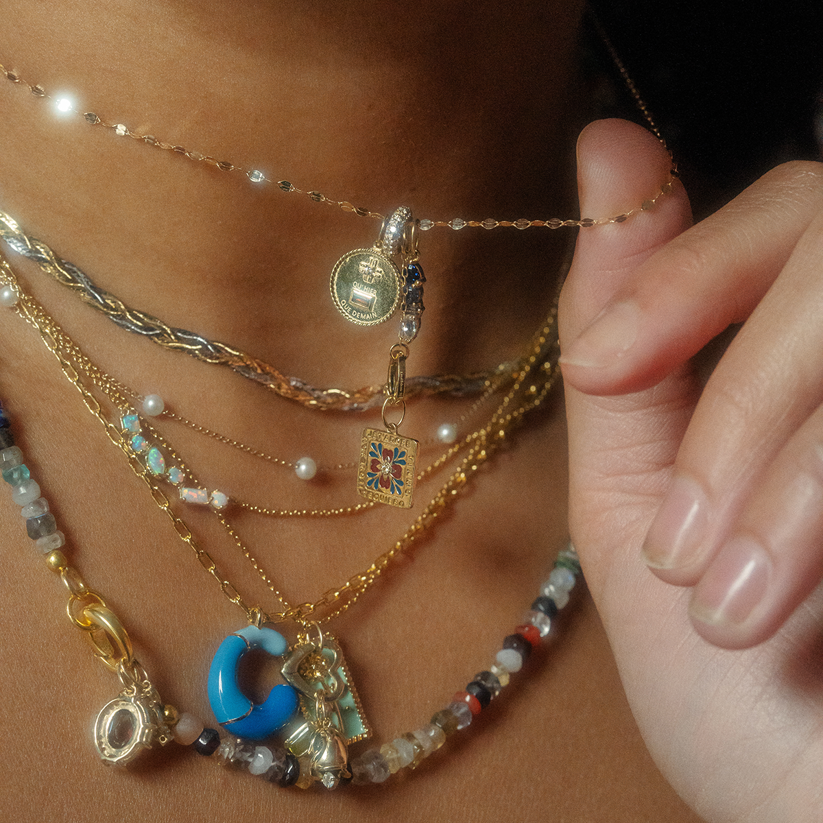 A close-up captures a person’s neck adorned with layered necklaces, including Solid Gold Statement Charm pendants, colorful beads, and a 9k yellow gold pearl and blue topaz pendant. A manicured hand gently holds one necklace to highlight its detail.