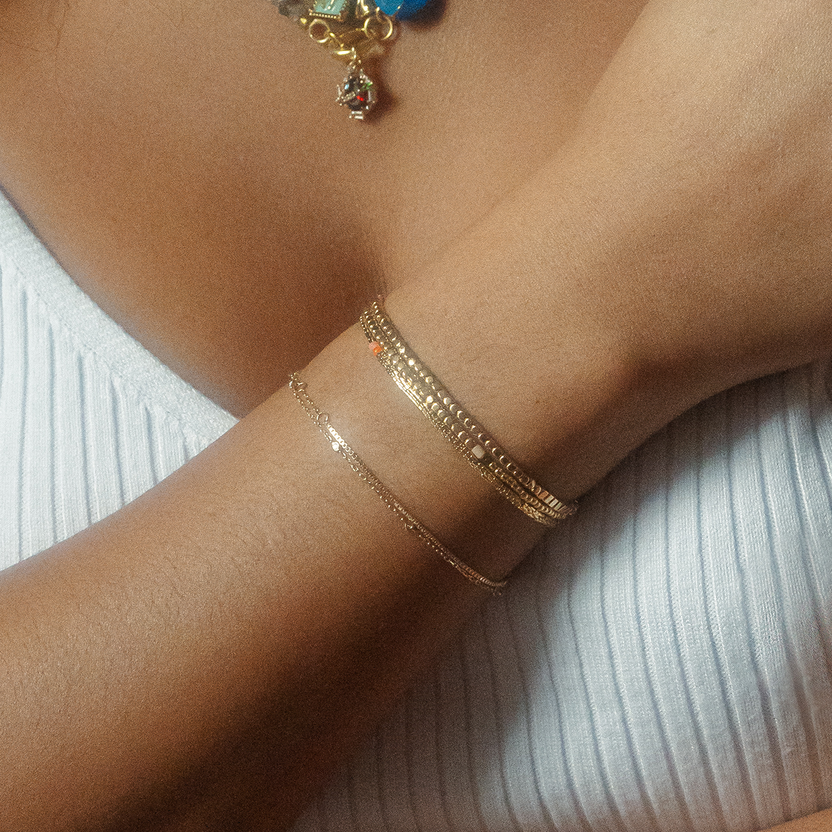 A close-up of a person wearing three gold bracelets, including the Solid Gold Simple Diamond Chain Bracelet, on their wrist with a white textured top. Part of a colorful necklace is also visible near the neckline.