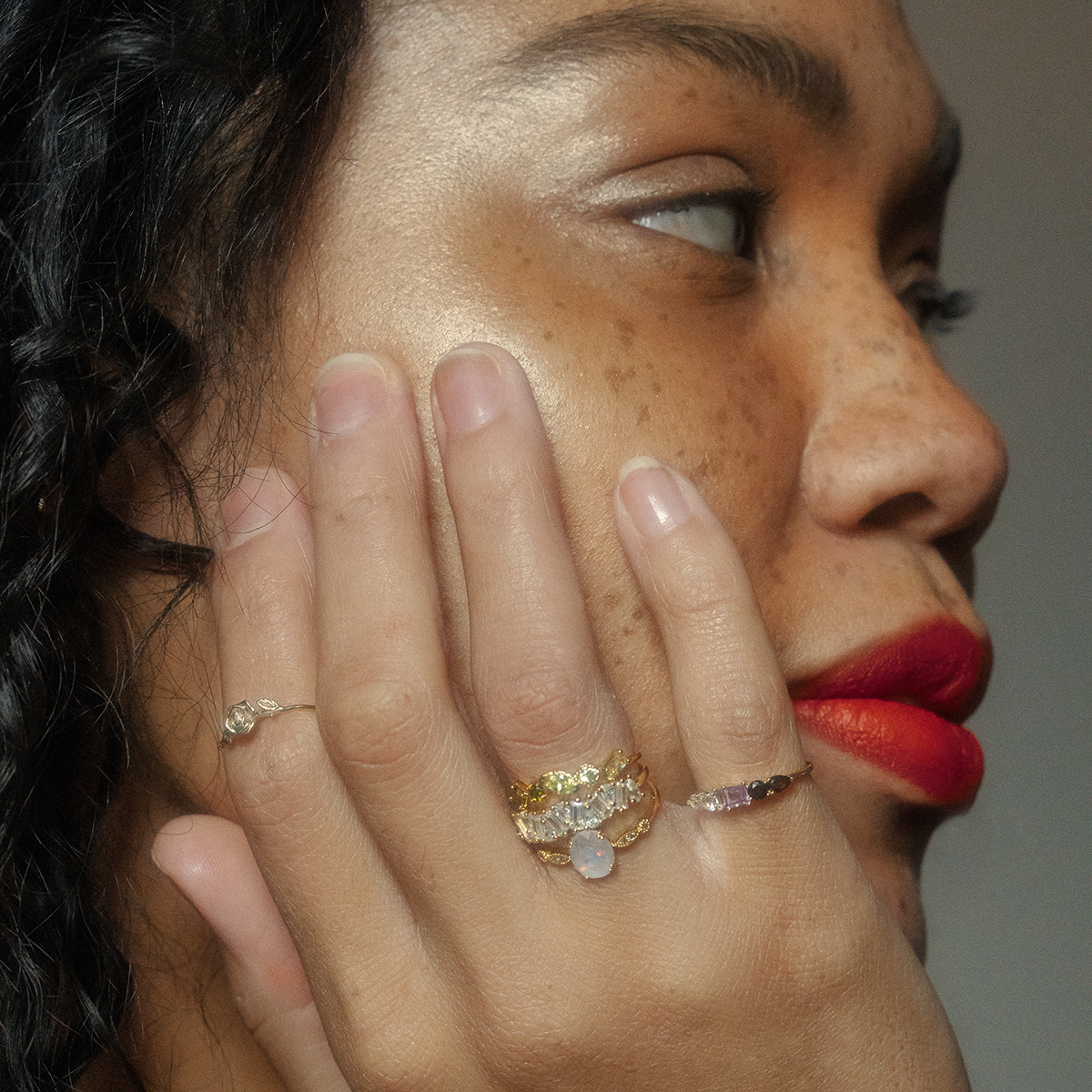 A woman with red lipstick and freckles rests her hand on her cheek, showing off a Solid Gold Birth Flower Ring among other gemstone rings. Her nails are natural as she gazes to the side.
