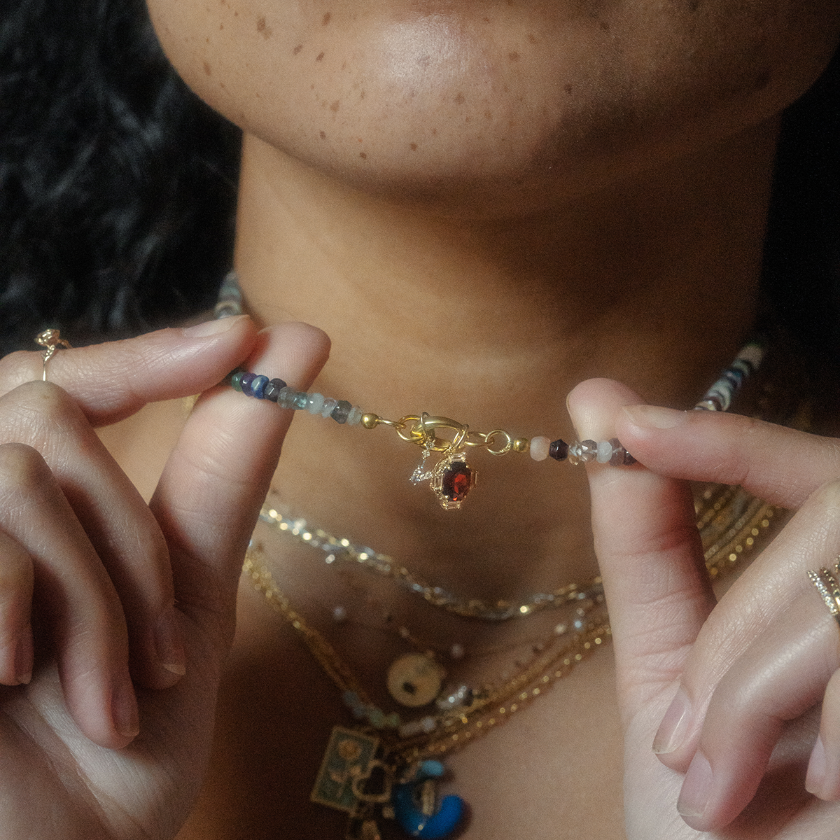 A person holds a beaded bracelet featuring the Solid Gold Vintage Inspired Birthstone Charm with a gold clasp, layered necklaces, and rings, while their softly visible lips and lower face appear in the background.