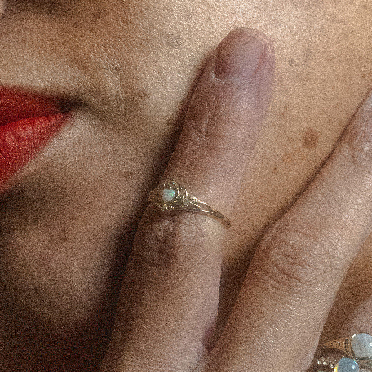 A close-up of a face with red lipstick and freckles, hand near cheek, wearing the Solid Gold Openwork Filigree Opal Heart Ring on their finger.
