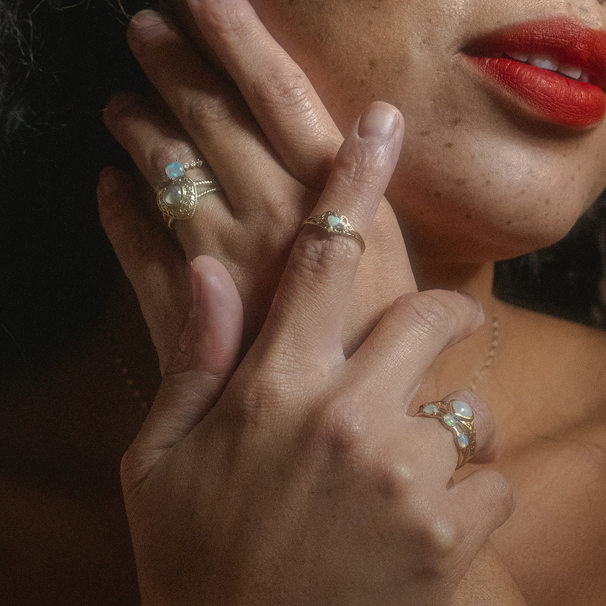 A close-up of a womans face and hands showcases bold red lipstick and sparkling gemstone rings, including the stunning Solid Gold Lovely Opal Ring. Soft lighting highlights her jewelry and smooth complexion.