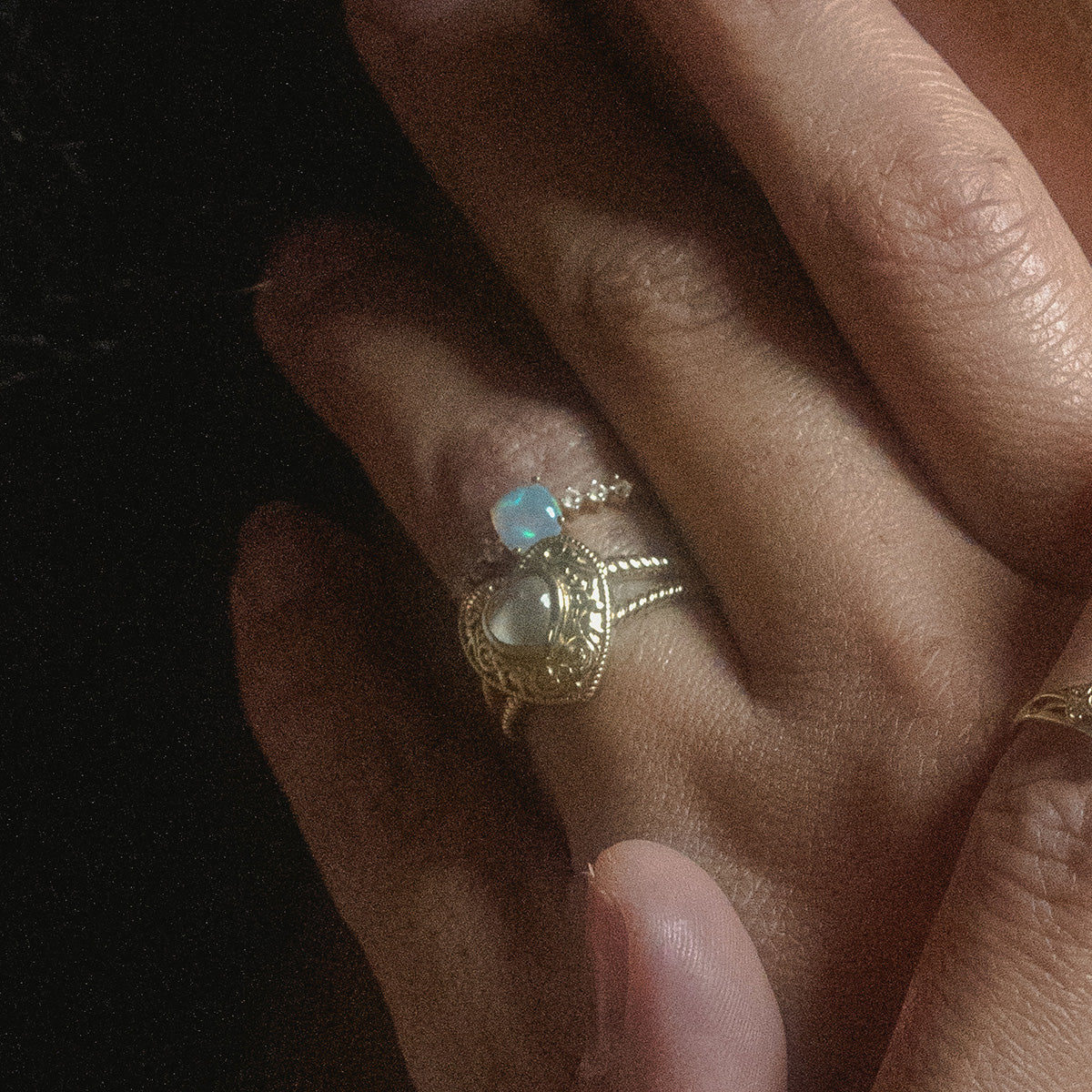 A close-up of hands, one resting atop the other, showcases two gold rings—one featuring the Solid Gold Rainbow Moonstone Heart Locket Ring. The lighting is soft and the background is dark.