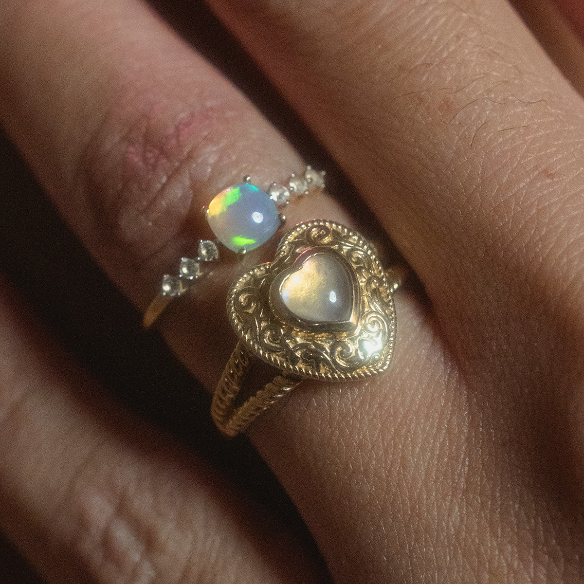 A person wears two gold rings: the Solid Gold Lovely Opal Ring, glowing with small diamonds, and another gold band with a heart-shaped gemstone set in ornate metalwork.