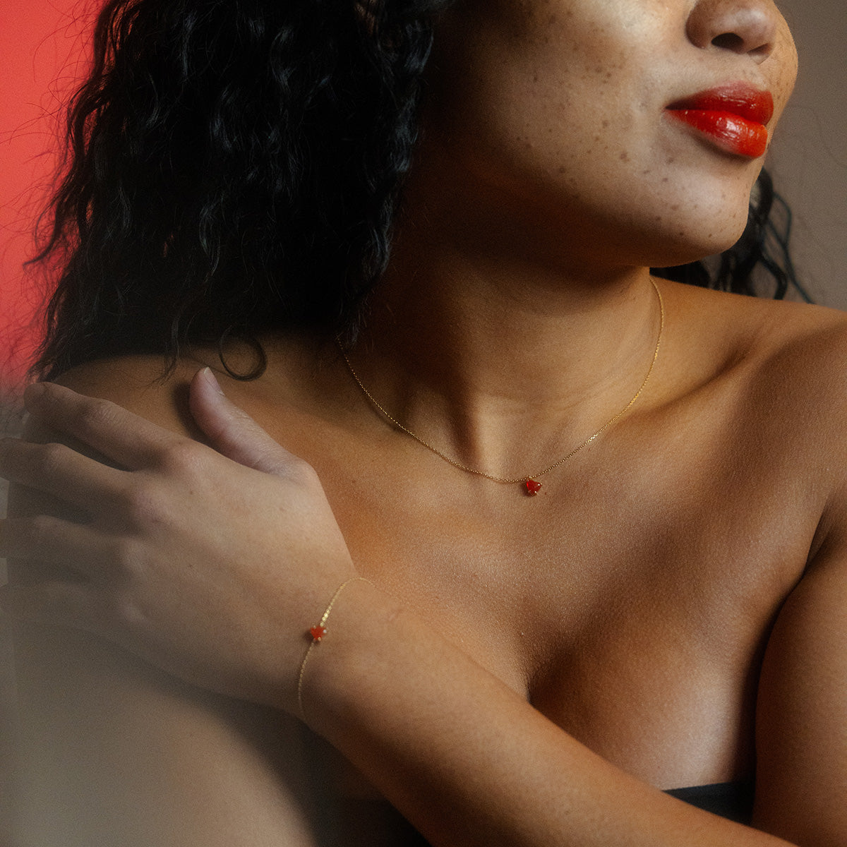 A woman with red lipstick wears the Limited Edition Full Heart Necklace Sample, a delicate 14k gold plated piece adorned with a small red flower charm, her hand resting on her shoulder as soft light creates a warm, intimate mood.