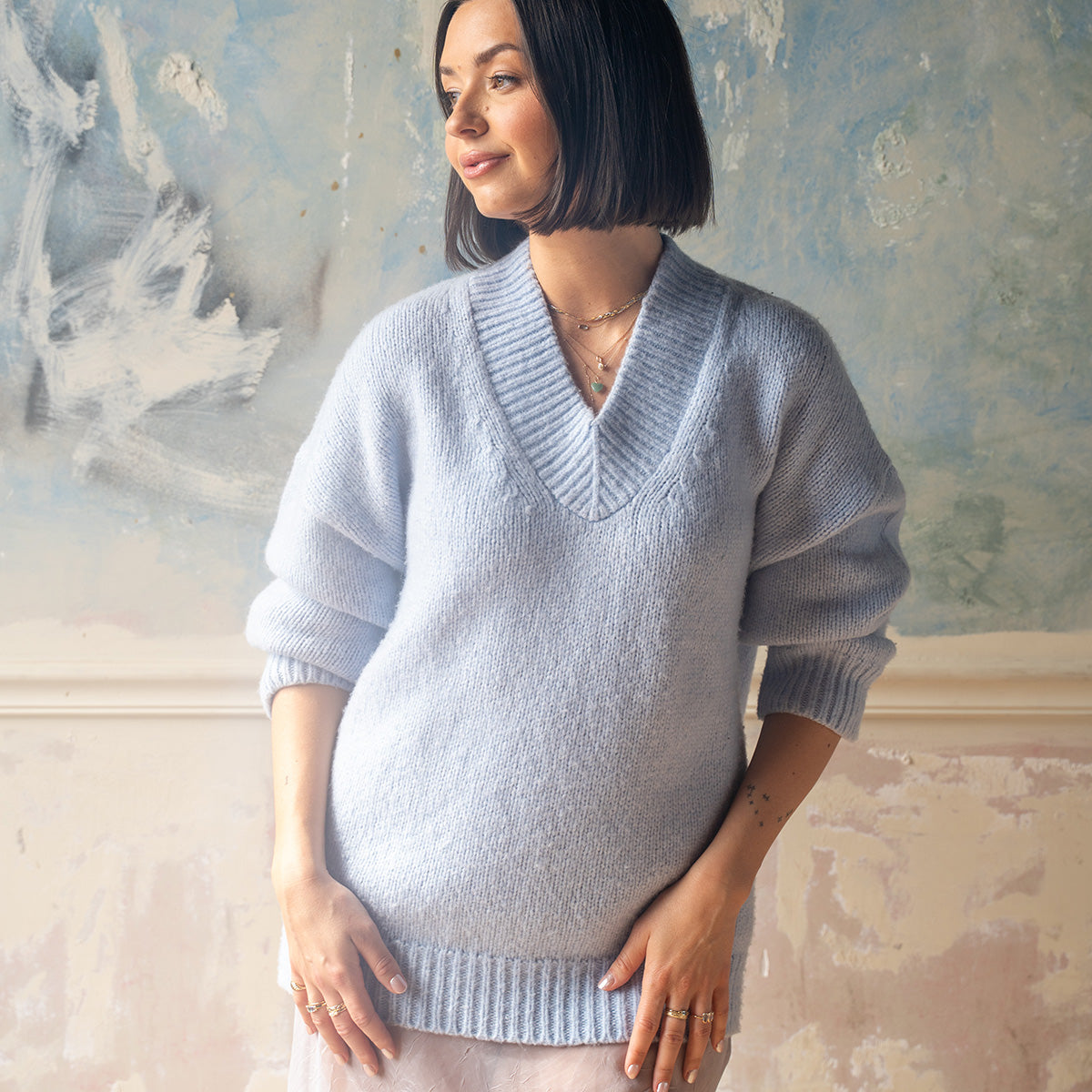 A woman with a bob haircut, in a light blue knit sweater and skirt, stands before a pastel wall, smiling softly. Her Solid Gold First Light Pastel Gemstone Ring adds a subtle touch of elegance as she poses with her hands by her waist.