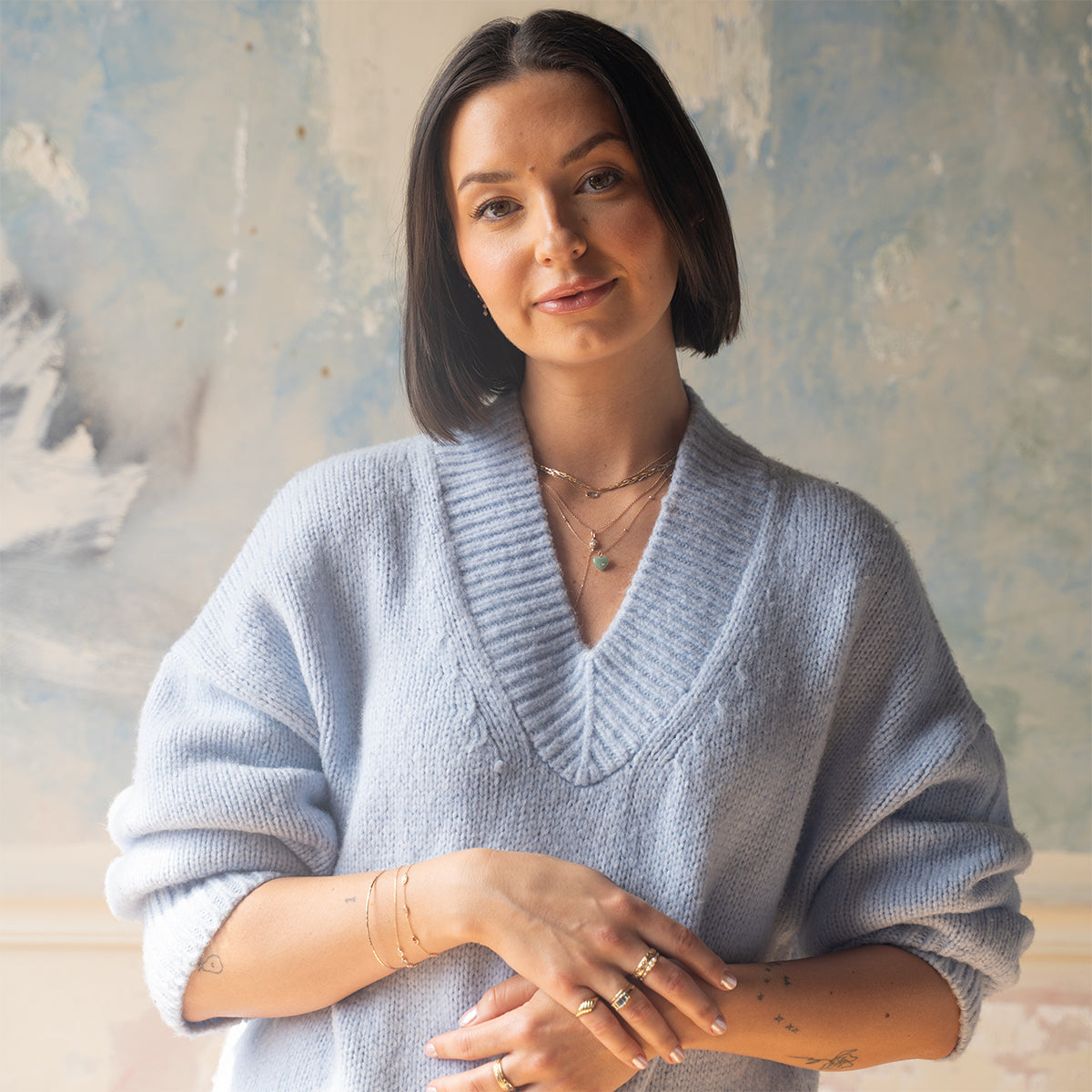A woman with short dark hair, wearing a light blue knit sweater and layered necklaces, stands indoors with hands gently crossed. She smiles softly while showcasing the Solid Gold Daybreak Moonstone and Blue Topaz Bracelet.