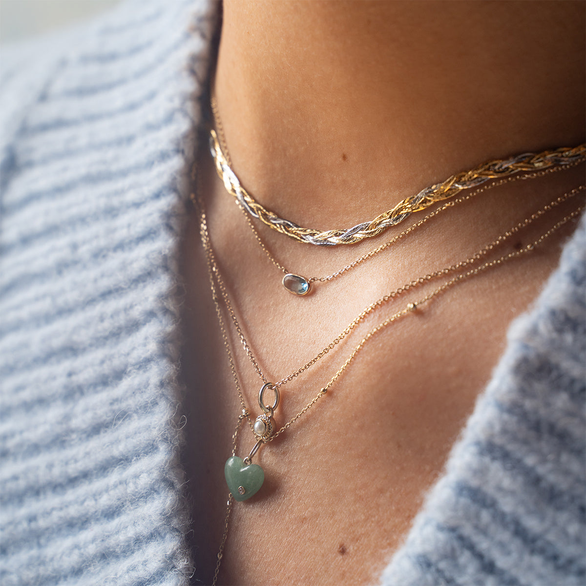 Close-up of a person in a blue knitted sweater wearing four layered gold necklaces, including a green heart-shaped pendant, a small round gemstone pendant, and the Solid Gold Daybreak Swiss Blue Topaz Necklace from Family Gold.