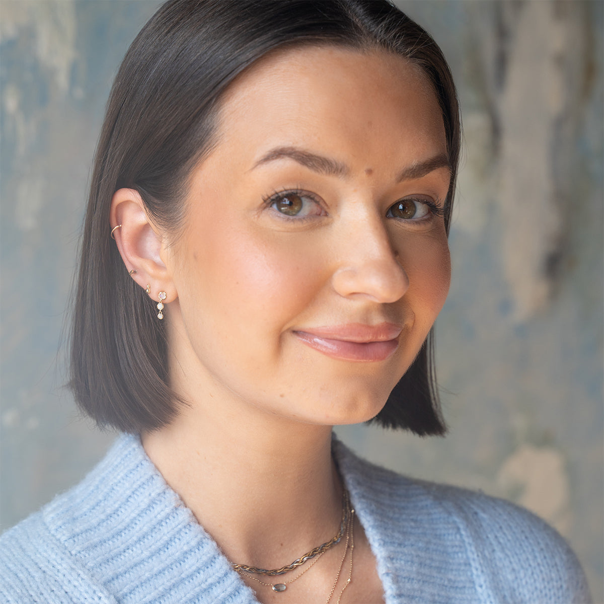 A woman with straight, dark brown bobbed hair wears a light blue sweater, small hoop earrings, and the Solid Gold Daybreak Swiss Blue Topaz Necklace. She smiles softly, facing slightly to the side against a blurred background.