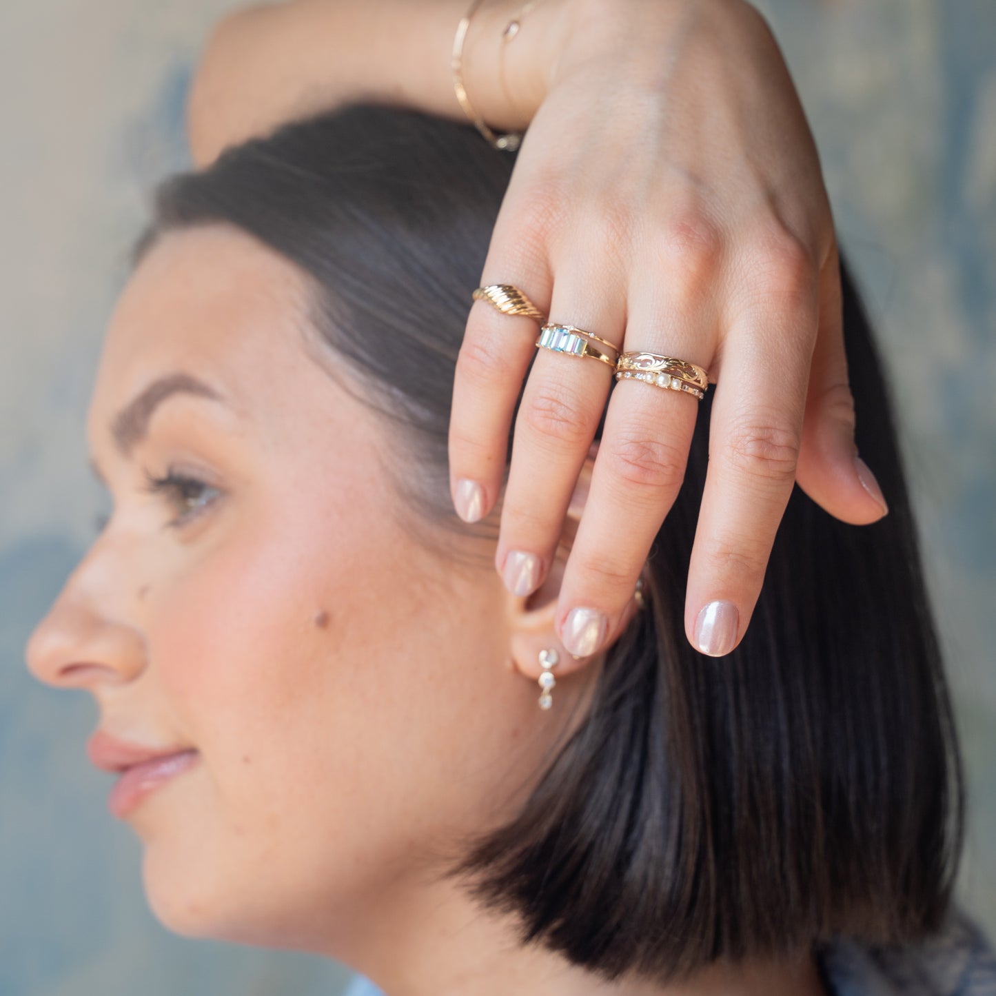 A woman with short dark hair poses, highlighting the Solid Gold Luminous Pearl Trio Ring alongside gold rings and a delicate bracelet. She wears a pearl earring and natural nude-toned nail polish.