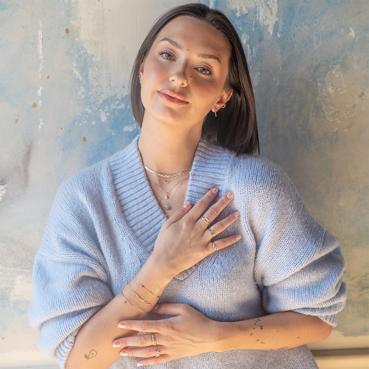 A woman with straight dark hair, wearing a light blue sweater, smiles gently in front of a textured wall, showcasing rings, bracelets, and the Family Gold Solid Gold Daybreak Swiss Blue Topaz Necklace.