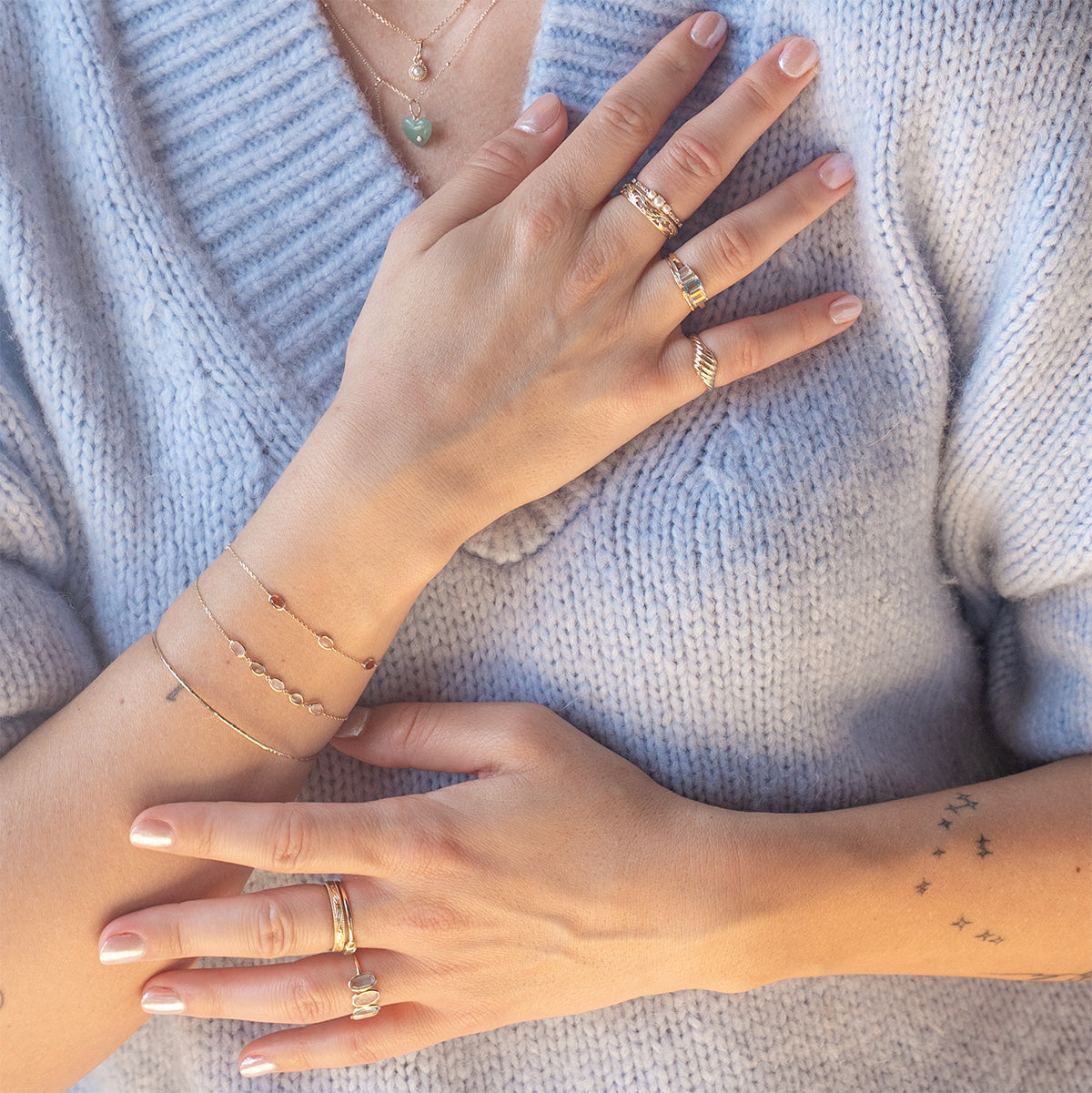 A person in a soft, light blue sweater crosses their hands over their chest, showcasing delicate bracelets, layered necklaces, dainty star tattoos, and the Solid Gold Luminous Pearl Trio Ring among other rings.