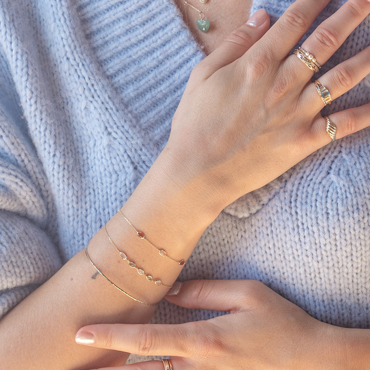 A person in a pale blue knitted sweater shows their hands adorned with delicate gold rings and the Solid Gold Daybreak Moonstone and Blue Topaz Bracelet, along with a necklace featuring a green heart-shaped pendant.