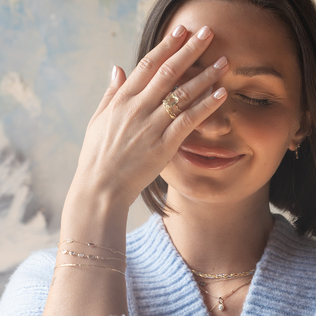 A woman with short brown hair smiles softly, covering one eye with her hand. She wears the Solid Gold Dewdrop Green Amethyst Statement Ring, paired with delicate bracelets and layered necklaces, and is dressed in a light blue textured top.