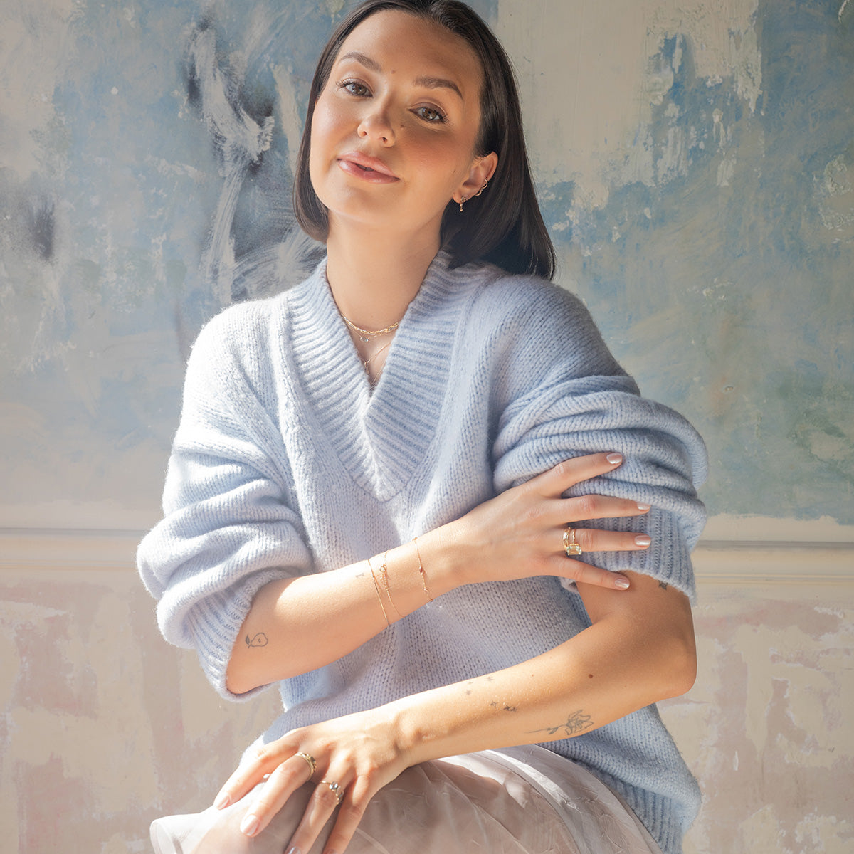 A woman with straight brown hair and subtle makeup sits against a pastel-painted wall, wearing a light blue sweater, white skirt, delicate jewelry, and the Solid Gold Dewdrop Green Amethyst Statement Ring on her tattooed fingers.