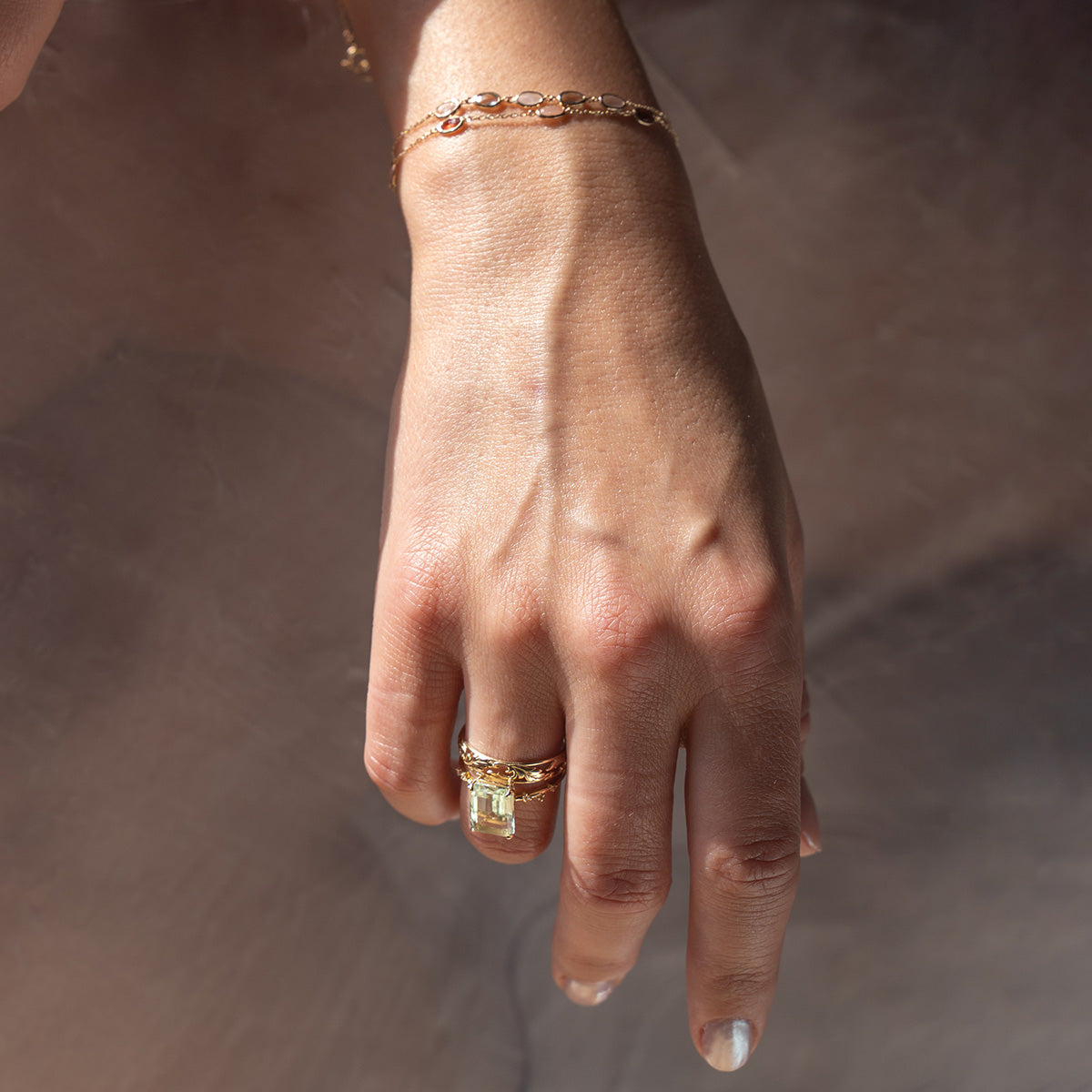 A hand with light skin tone wears a solid gold bracelet and two gold rings, including the Solid Gold Dewdrop Green Amethyst Statement Ring with a large rectangular gemstone, against a soft neutral background. The nails are short and natural.