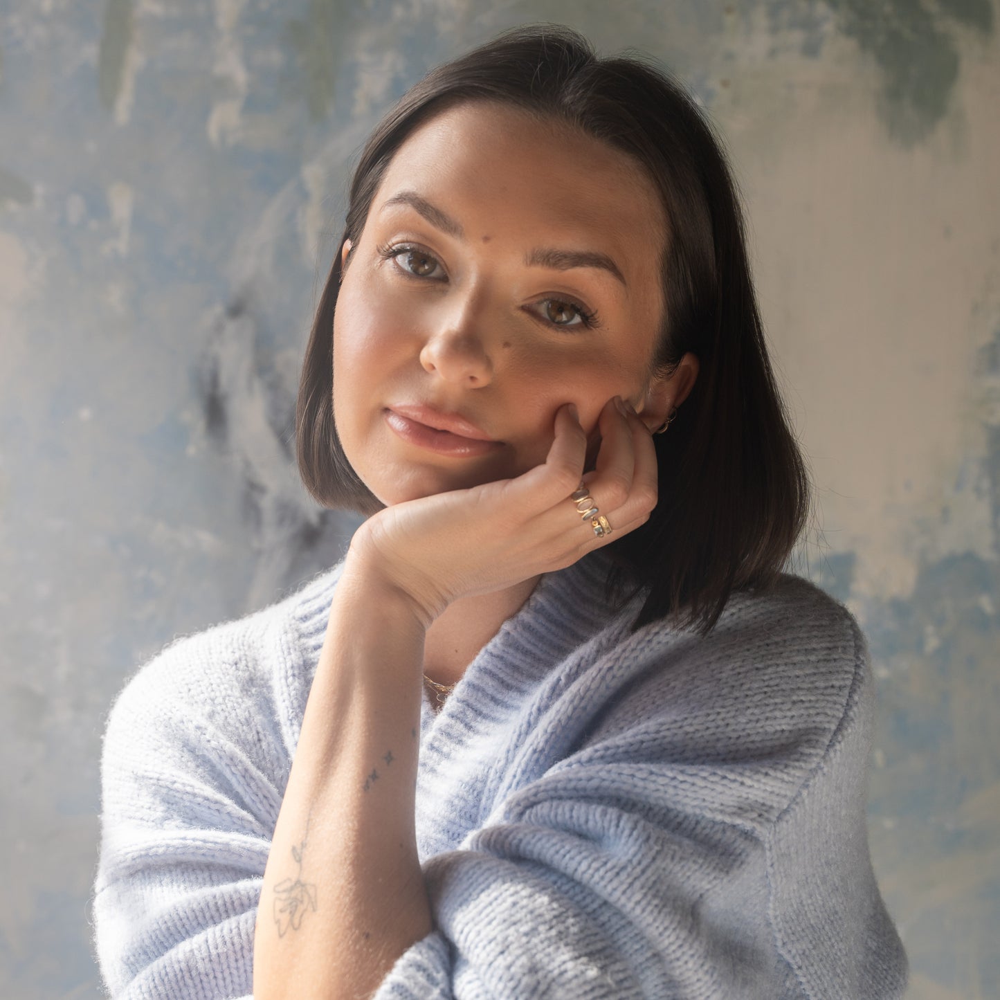 A woman with straight dark hair in a light sweater thoughtfully gazes at the camera, her face softly lit by natural light. She wears the Solid Gold Daybreak Moonstone and Blue Topaz Ring, which subtly complements the tattoo on her wrist.