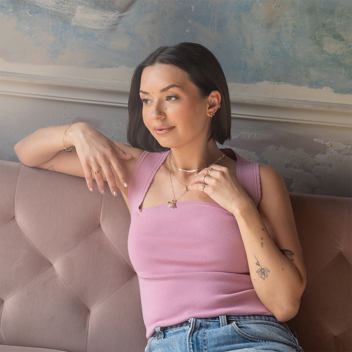 A woman with dark hair and tattoos sits on a pink sofa in a sleeveless top and jeans, wearing several rings, bracelets, and the Solid Gold Luminous Pearl Trio Necklace layered with other pieces as she looks thoughtfully to the side.
