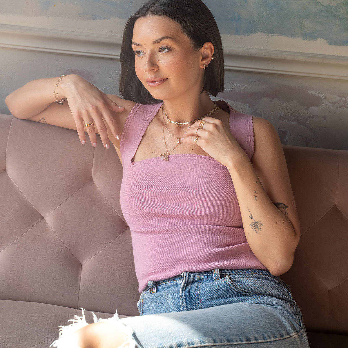 A woman with short brown hair sits on a pink sofa, wearing a sleeveless pink top, ripped blue jeans, and layered necklaces. She wears Solid Gold Soft Glow Pearl and Moonstone Drop Studs and rings, gazing thoughtfully to the side.