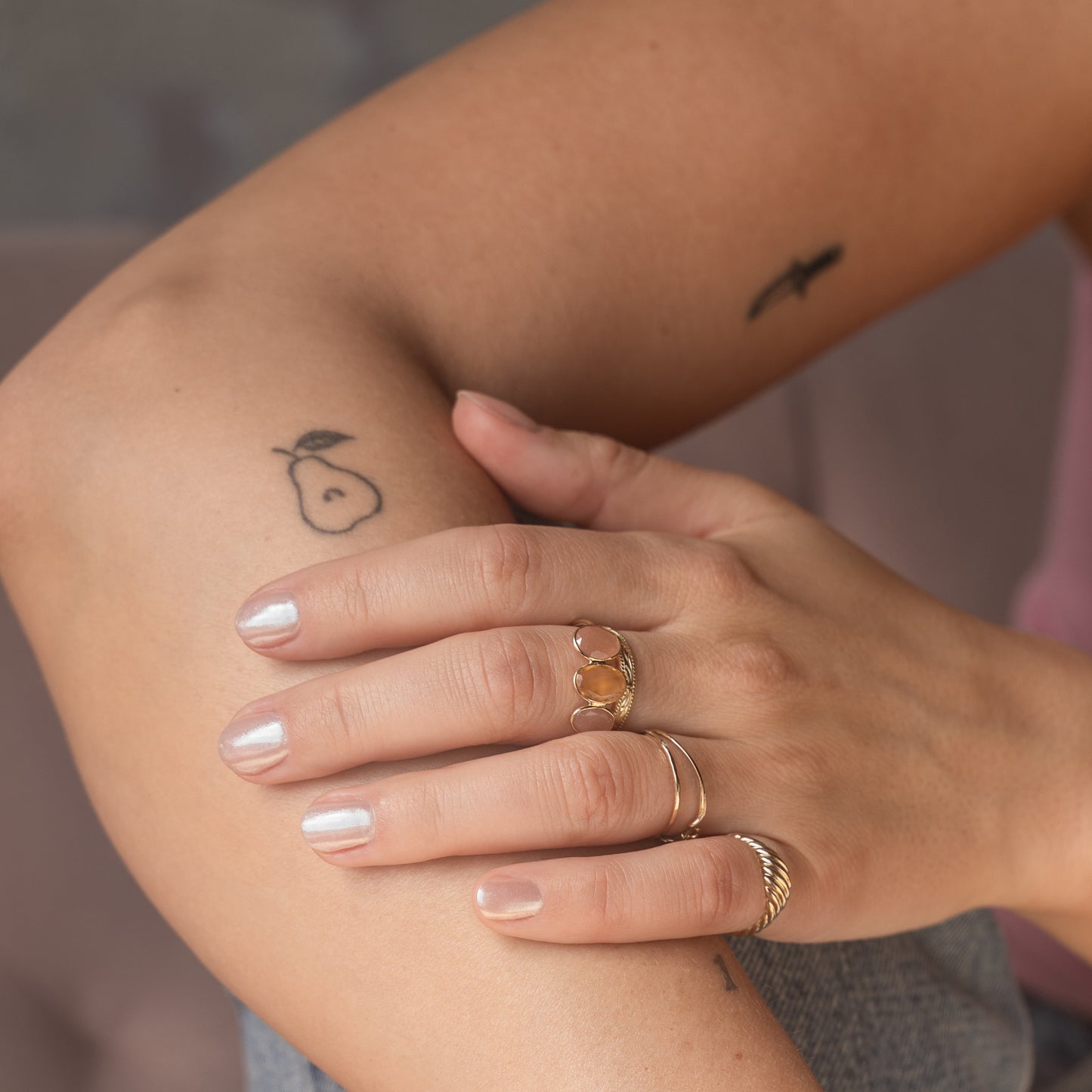 A close-up of hands with light pink nail polish showcases several Family Gold exclusive solid gold rings, including the Solid Gold Morning Light Citrine and Sunstone Ring, plus a small pear tattoo on the inner arm and another partially visible upper arm tattoo.