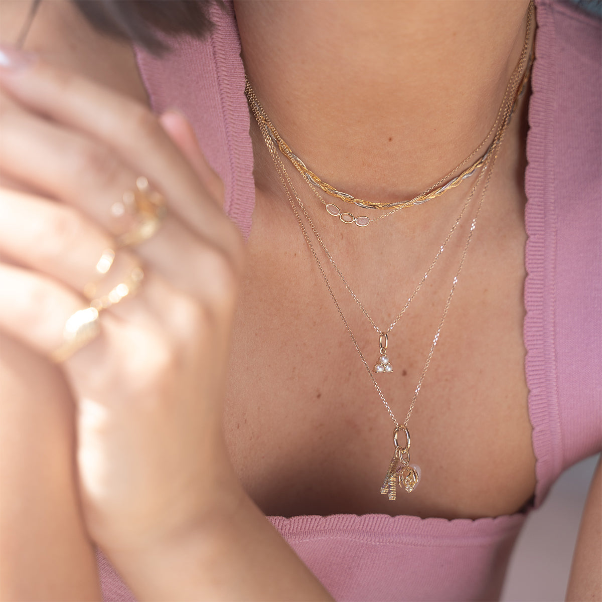 A woman in a scalloped-edge pink top showcases the Solid Gold Luminous Pearl Trio Necklace layered with silver pieces and gold rings. Her face is partially out of frame.