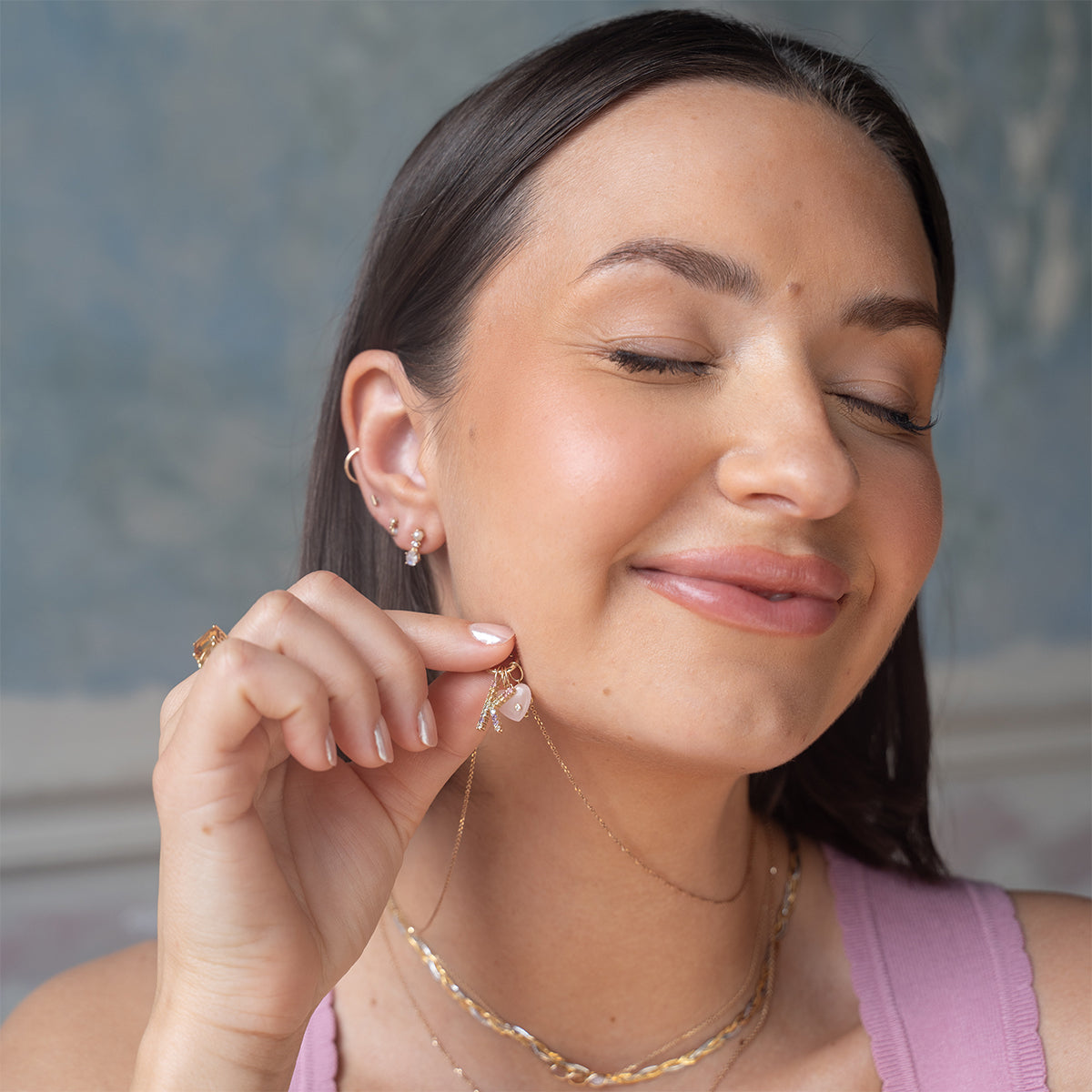 A woman with long dark hair smiles with her eyes closed, holding the Solid Gold Full Heart Crystal Charm necklace near her face. She wears multiple earrings, a pink top, and layered gold necklaces.