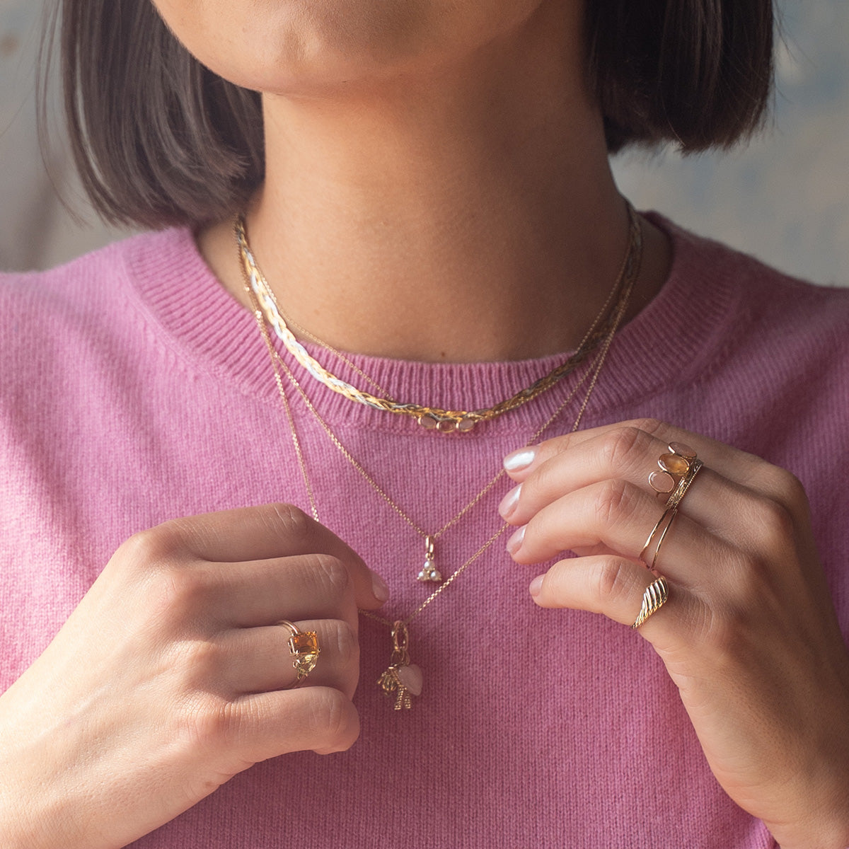 A woman in a pink sweater wears the Solid Gold Luminous Pearl Trio Necklace, layered with gold jewelry. Her short dark hair and light pink manicure accent her look, as her partially visible face adds elegance to the scene.