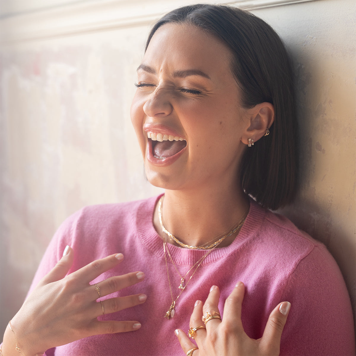 A woman with straight dark hair, in a pink sweater and gold jewelry featuring the Solid Gold Luminous Pearl Trio Necklace, laughs joyfully with her eyes closed as she leans against a light-colored wall.