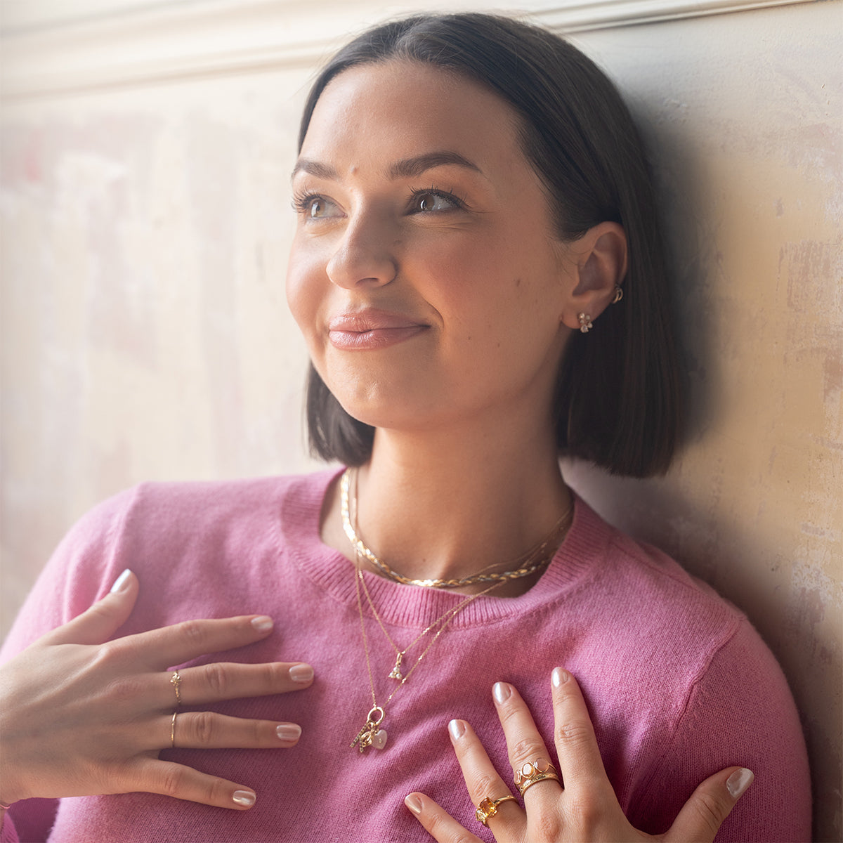 A woman with straight dark hair in a pink sweater smiles softly and looks upward. She layers multiple gold necklaces, including the Solid Gold Full Heart Crystal Charm, along with gold rings and earrings, her hands gently touching her chest.